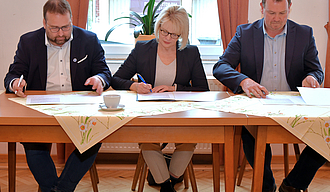 Drei Personen sitzen an einem Tisch und unterzeichnen jeweils ein Exemplar der Absichtserklärung